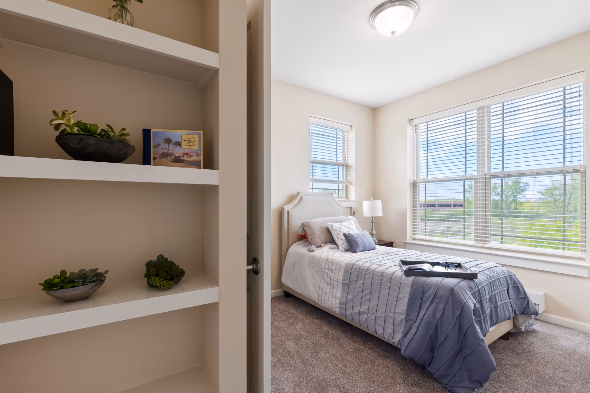 A bright bedroom with a single bed covered in a gray and white striped comforter and multiple pillows. There is a tray with a book and glasses on the bed. The room has large windows with blinds letting in natural light, a bedside table with a lamp, and beige walls. To the left, there is a built-in white shelf with small potted plants and a book.