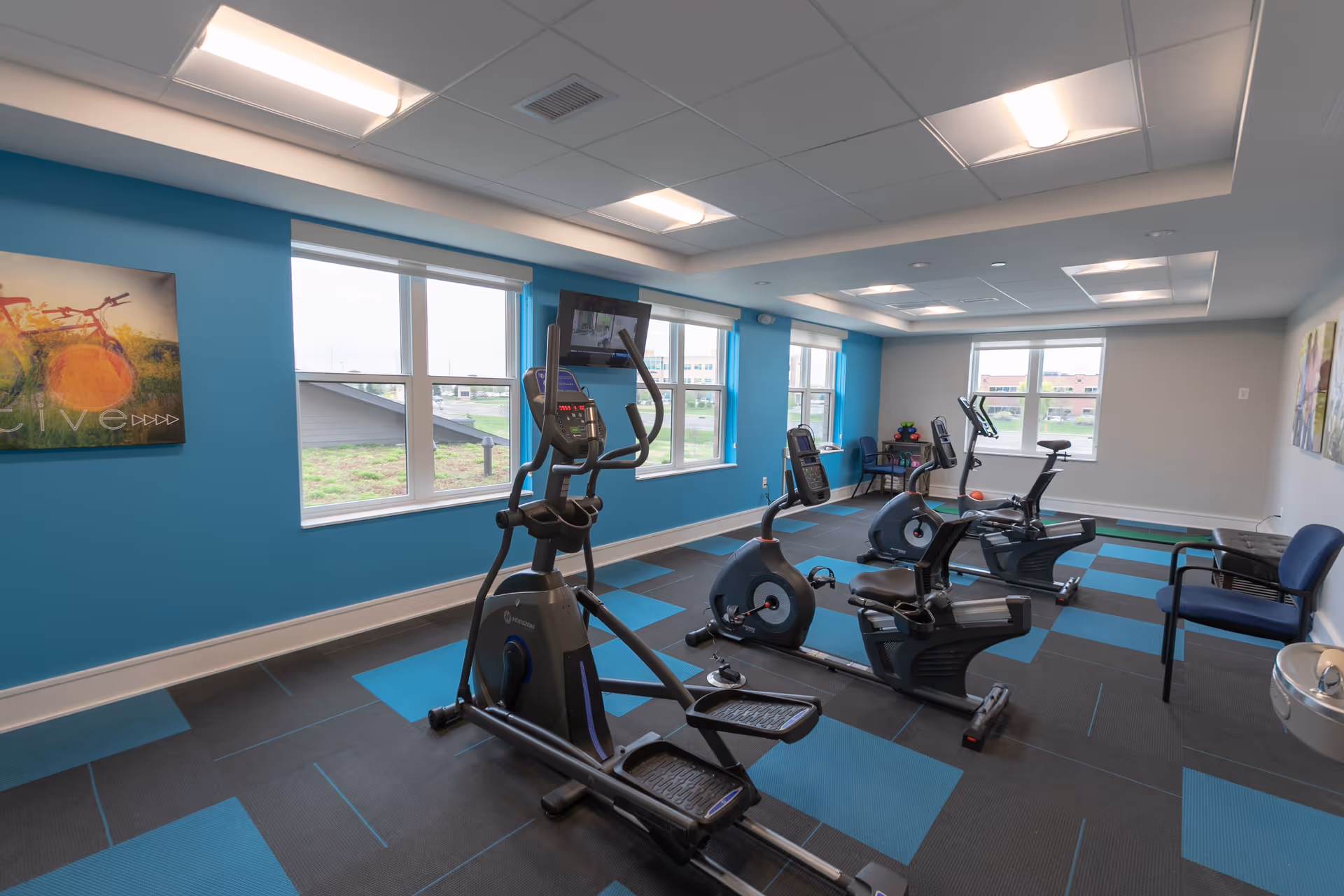 A fitness room with exercise equipment including an elliptical machine and two recumbent bikes. The room has large windows letting in natural light, blue and gray checkered flooring, blue walls, and ceiling lights. There are chairs and a water fountain along the walls, and a colorful painting of a bicycle on one wall.