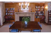 A cozy room with a wooden table and six chairs around it, a potted plant centerpiece on the table, a fireplace with a mirror above it, two bookshelves filled with books on either side of the fireplace, and two chairs placed next to the bookshelves. The room has warm lighting from a chandelier and wall sconces.