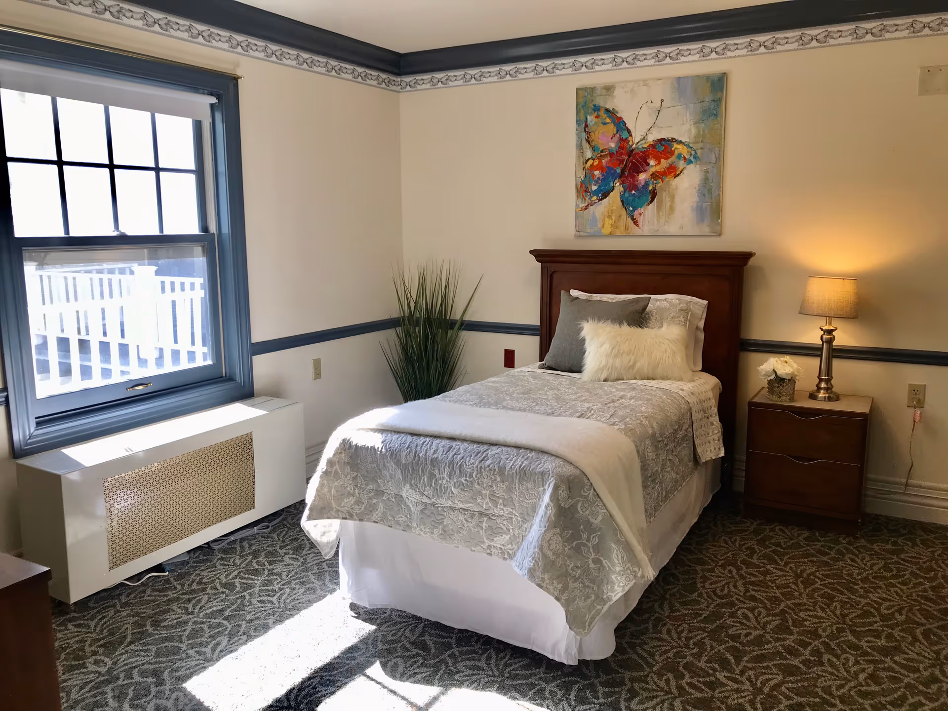 Sunlit single-occupancy bedroom with a neatly made twin bed, nightstand and lamp, window, and colorful butterfly artwork.