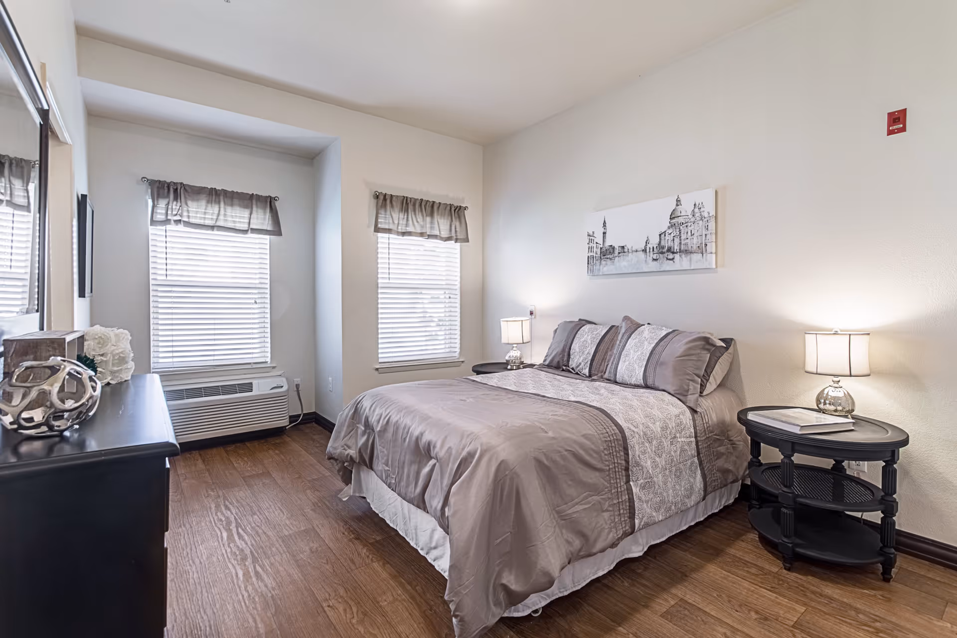 A neatly made bed with gray and white bedding in a bedroom with two windows covered by gray valances. There are two bedside tables with lamps on either side of the bed, a black dresser with decorative items, and a framed black and white cityscape artwork hanging above the bed. The room has wooden flooring and white walls.
