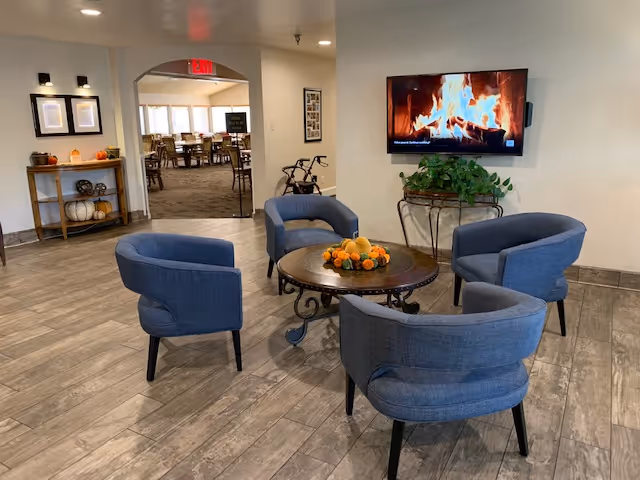 A cozy seating area with four blue upholstered chairs arranged around a round wooden table decorated with a fall-themed centerpiece. A flat-screen TV mounted on the wall displays a fireplace scene. In the background, there is an archway leading to a dining area with tables and chairs. The floor is covered with wood-look tiles, and a small console table with decorative pumpkins and framed pictures is visible near the archway.