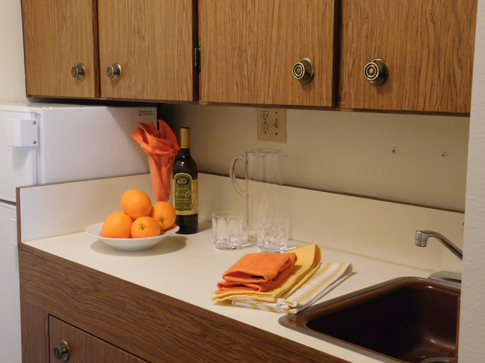 A kitchen countertop with a bowl of oranges, a bottle of olive oil, a glass pitcher, two glasses, and folded kitchen towels. Above the counter are wooden cabinets with round knobs, and to the right is a sink with a faucet.