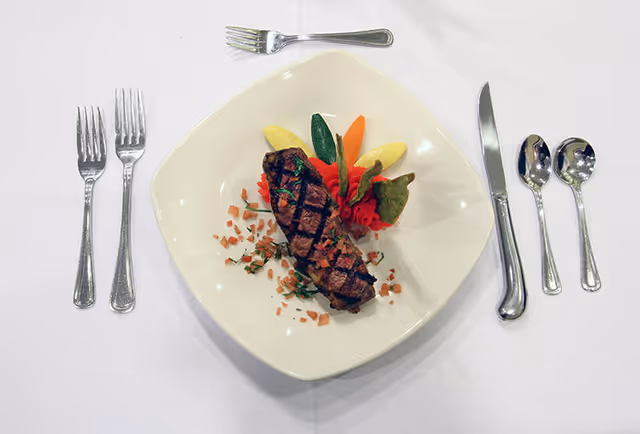 A plated meal featuring a grilled steak garnished with chopped herbs and small diced vegetables, accompanied by colorful vegetable garnishes on a white square plate. The plate is set on a white tablecloth with a formal place setting including three forks on the left, a knife and two spoons on the right, and a small fork above the plate.