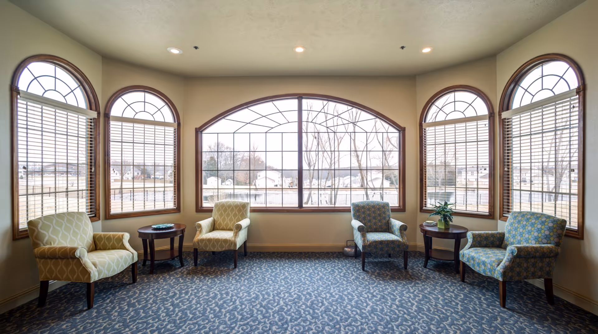 Sunlit common room with upholstered armchairs and side tables arranged before large arched windows overlooking the grounds.