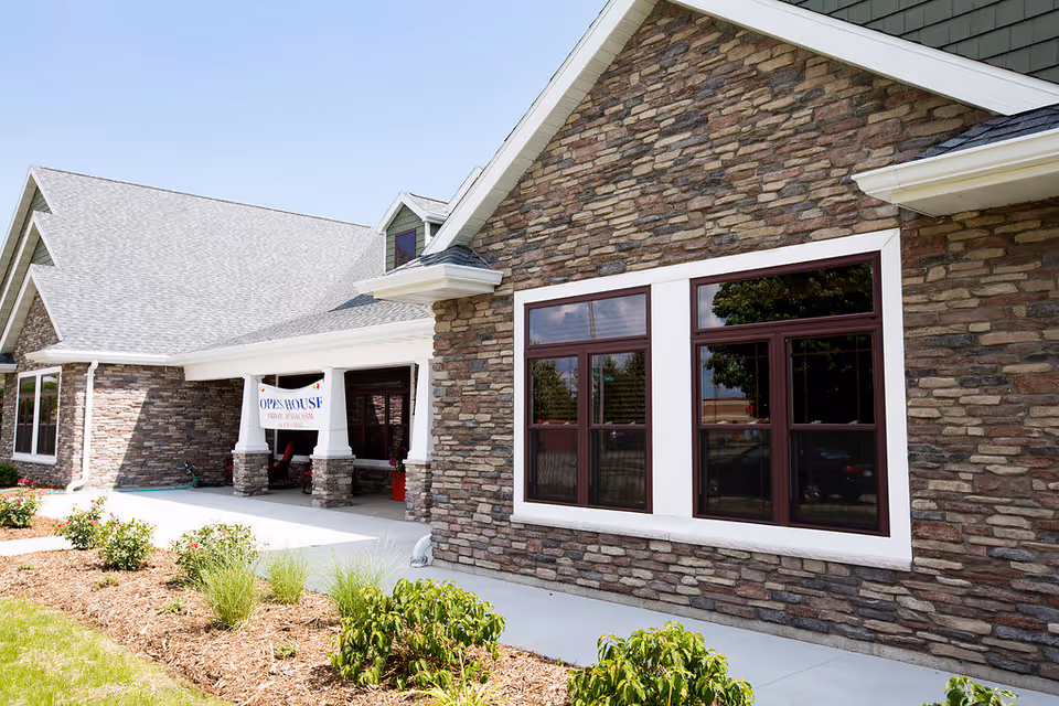 Stone-faced single-story assisted living facility front with large windows, a covered entrance and landscaped beds.