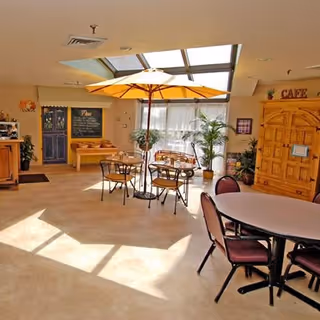 A bright indoor seating area with a large yellow umbrella over a round table with chairs. There are additional tables and chairs, potted plants, a wooden cabinet labeled 'CAFE', and a chalkboard menu on the wall. Skylights and large windows allow natural light to fill the space.