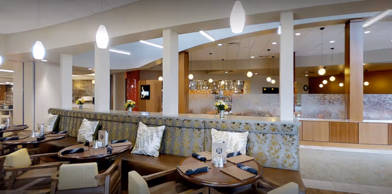 Dining room with round tables, cushioned banquette seating and decorative pillows under pendant lighting facing a service area.