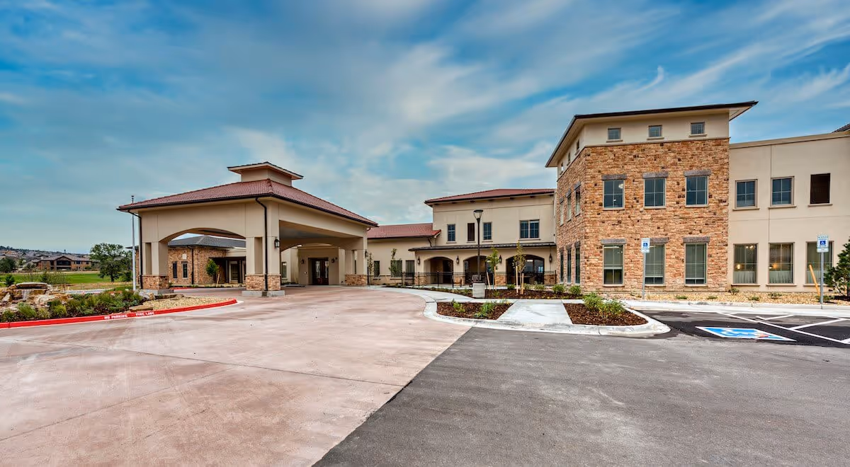Front exterior of a modern senior living facility with a covered porte-cochère, stone facade, landscaped beds, and a parking area under a partly cloudy sky.