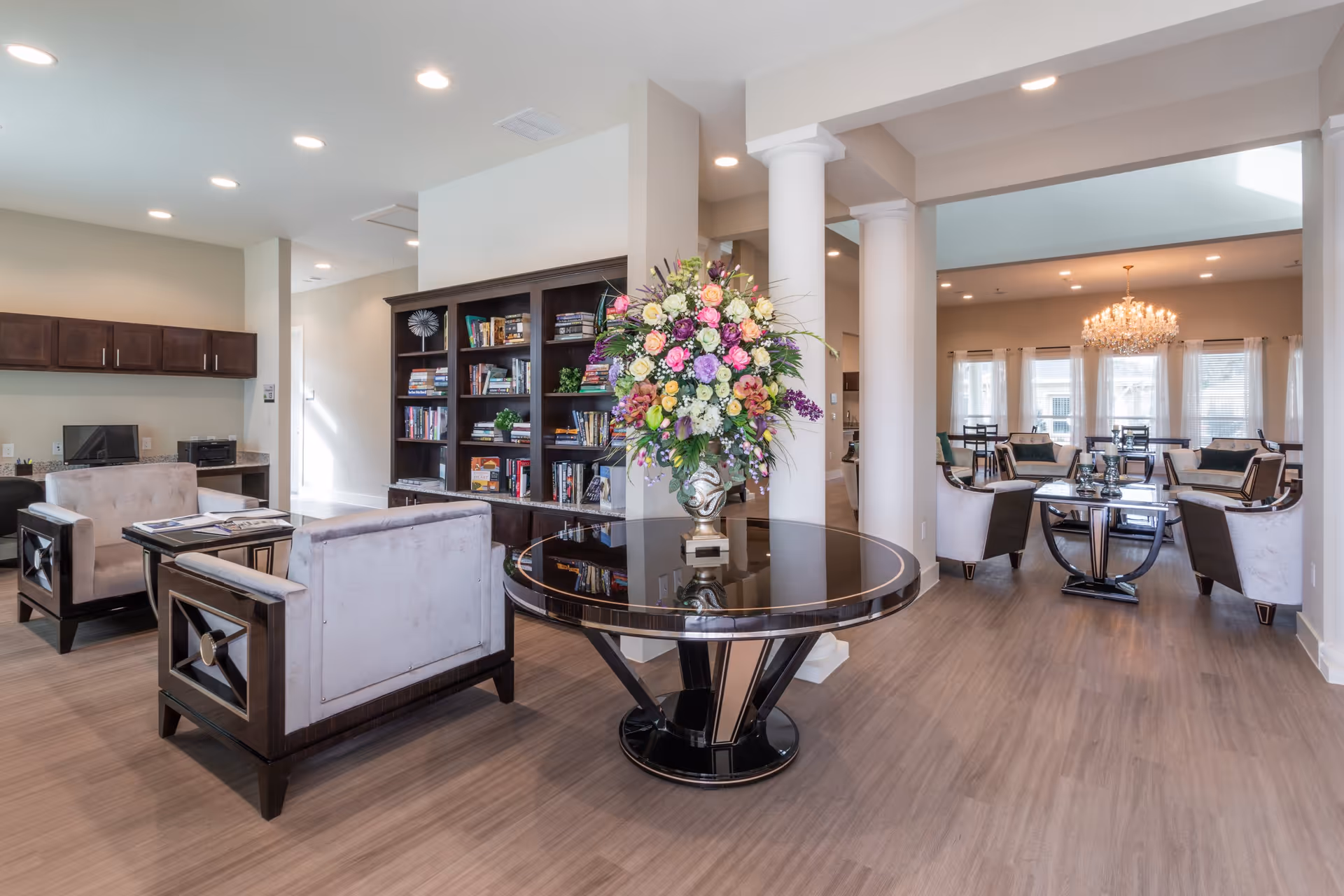 Spacious bright senior living lounge with seating areas, bookshelves, and a central table topped by a large floral arrangement.