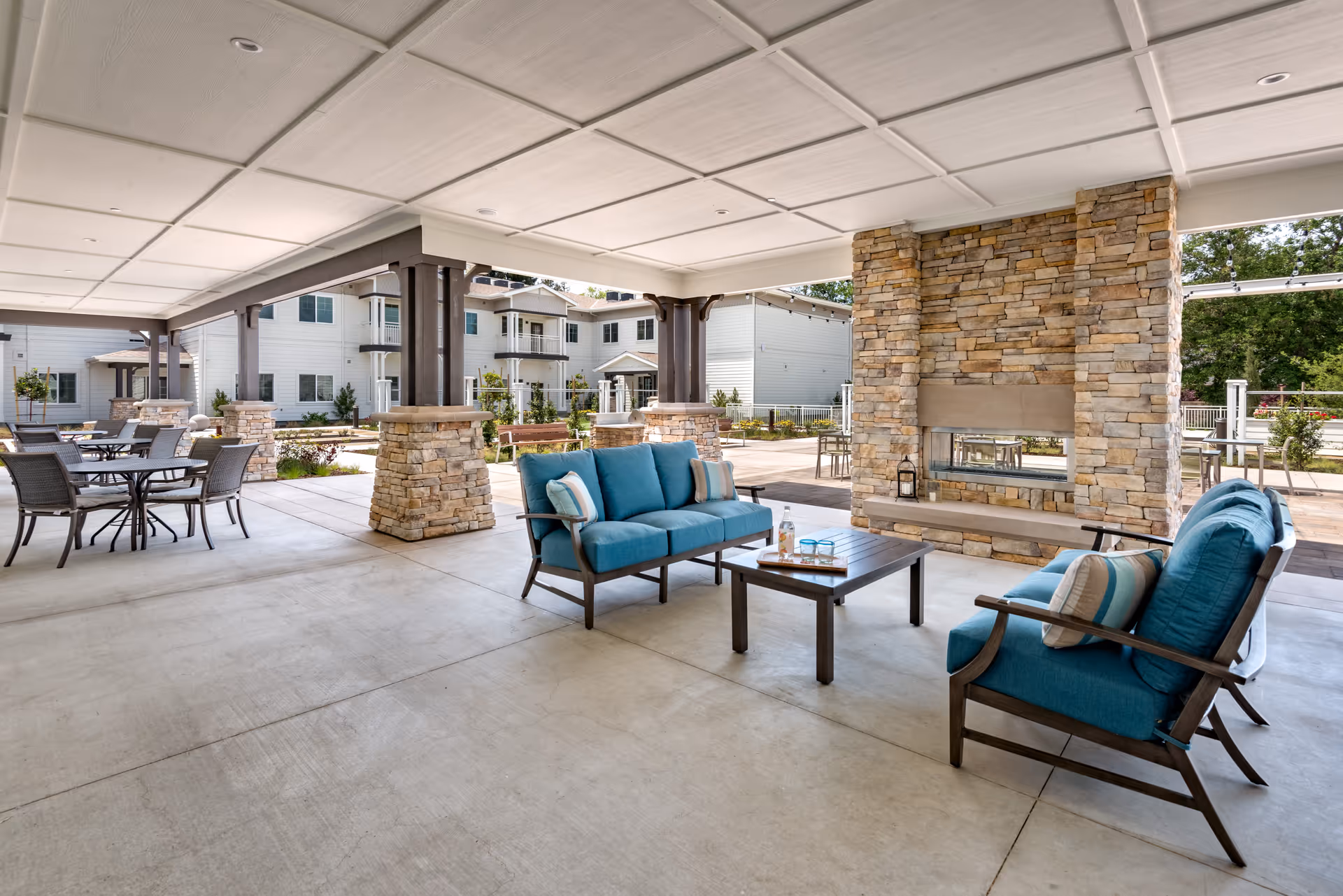 Covered outdoor seating area with blue cushioned chairs and a sofa around a wooden coffee table, next to a stone fireplace. Several round tables with chairs are visible in the background, along with a multi-story building and greenery.
