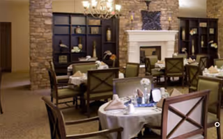 A cozy dining room with several round tables covered with white tablecloths, set with napkins and tableware. The room features a stone fireplace with a mantle and built-in dark wood shelves on either side. The walls are light-colored, and there is a chandelier hanging from the ceiling. The chairs have wooden frames with cushioned seats.