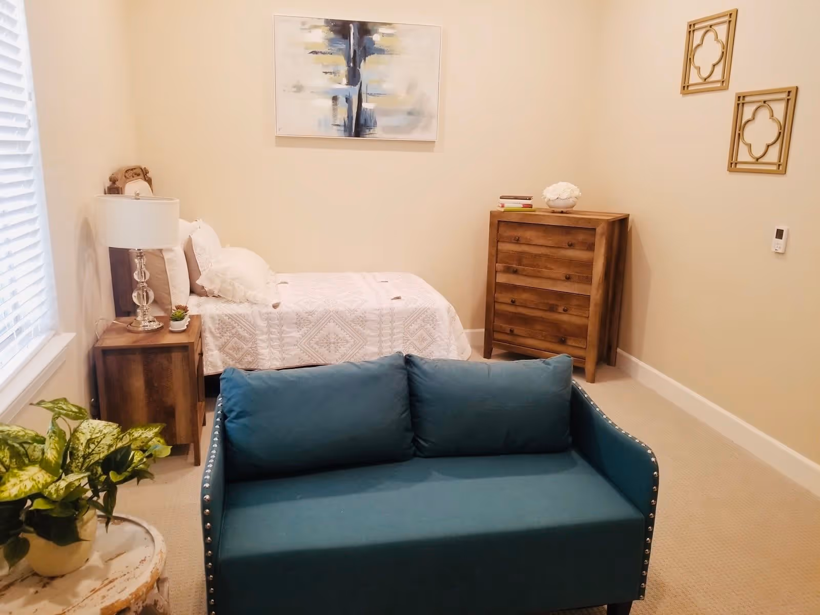 A cozy bedroom with a single bed covered in a white quilt, a wooden nightstand with a lamp and small plant, a wooden dresser with decorative items on top, a teal upholstered loveseat, a small round table with a potted plant, and abstract artwork on the wall.
