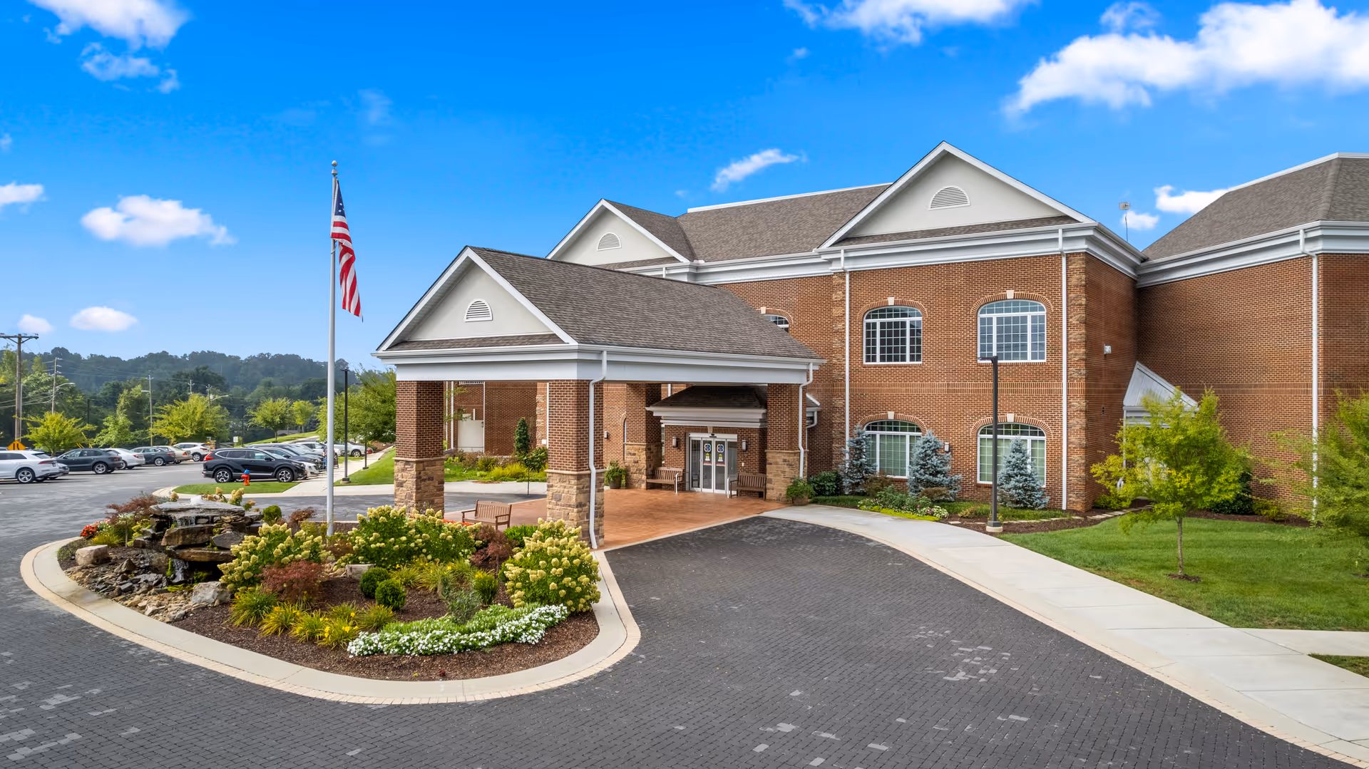 Front entrance of a brick senior living building with a covered porte-cochere, flagpole, landscaped roundabout and parked cars.