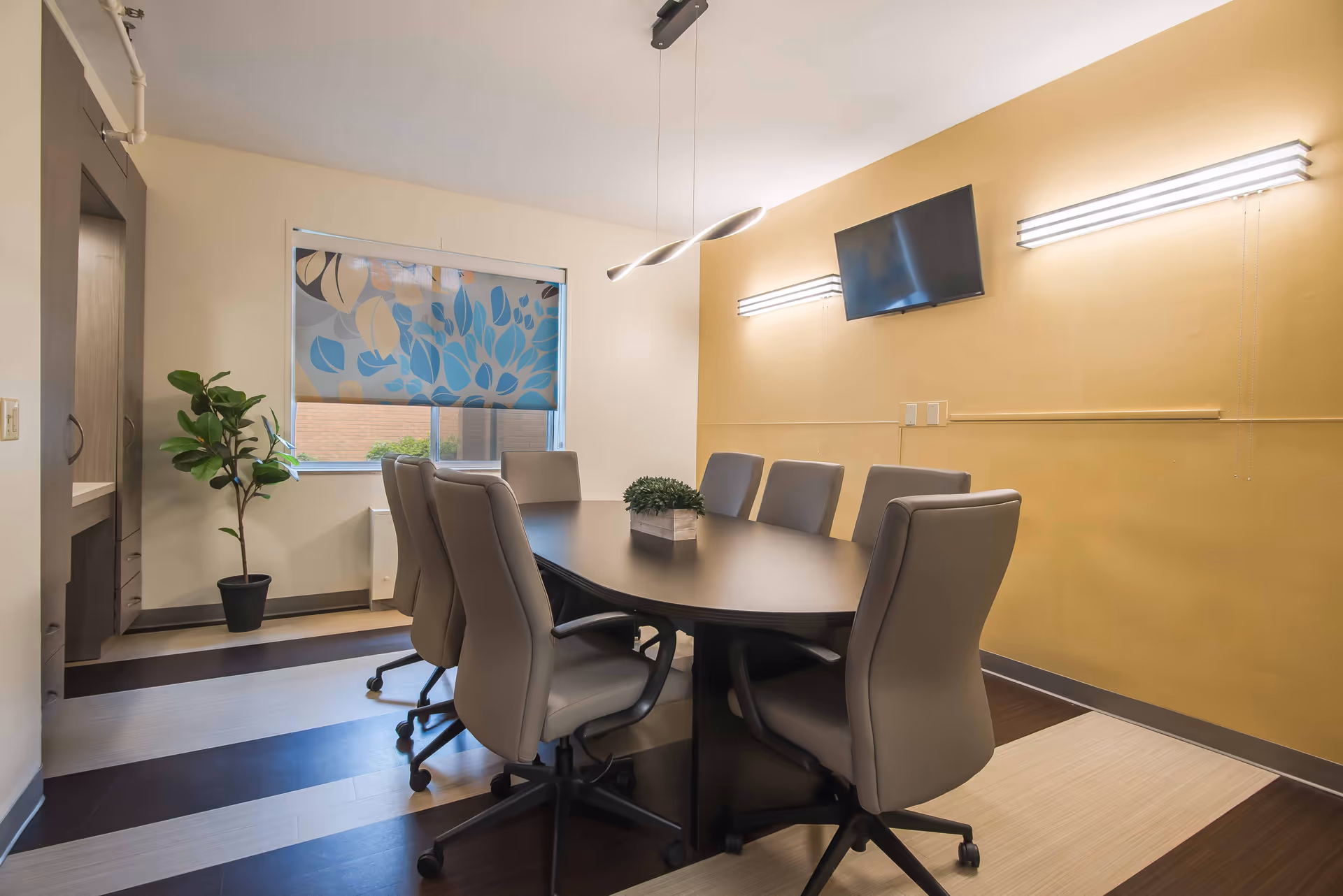 A modern conference room with a long oval table surrounded by eight gray swivel chairs. The room has a window with a decorative blue and beige leaf-patterned blind, a potted plant in the corner, a wall-mounted flat screen TV, and two horizontal wall lights on a yellow wall.