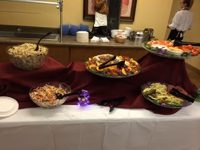 Buffet table with various dishes including a large bowl of salad, a platter of mixed vegetables, and other food items arranged on a burgundy cloth. A person in a white shirt and black apron stands in the background near the counter with plates and bowls stacked.