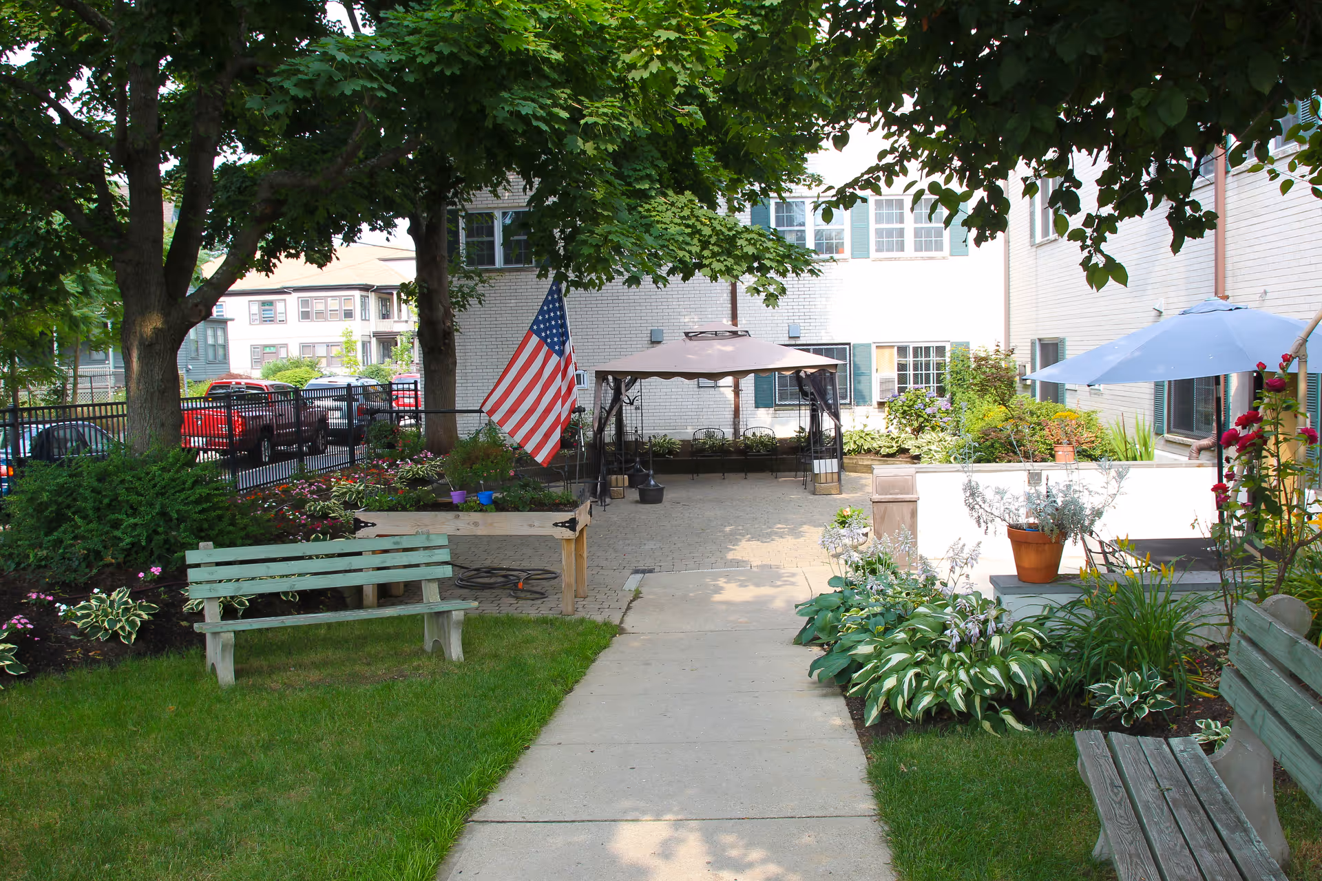 Outdoor courtyard area at Elmwood Nursing & Rehabilitation Center with green benches, a paved walkway, flower beds, an American flag, and shaded seating areas under umbrellas and a gazebo.