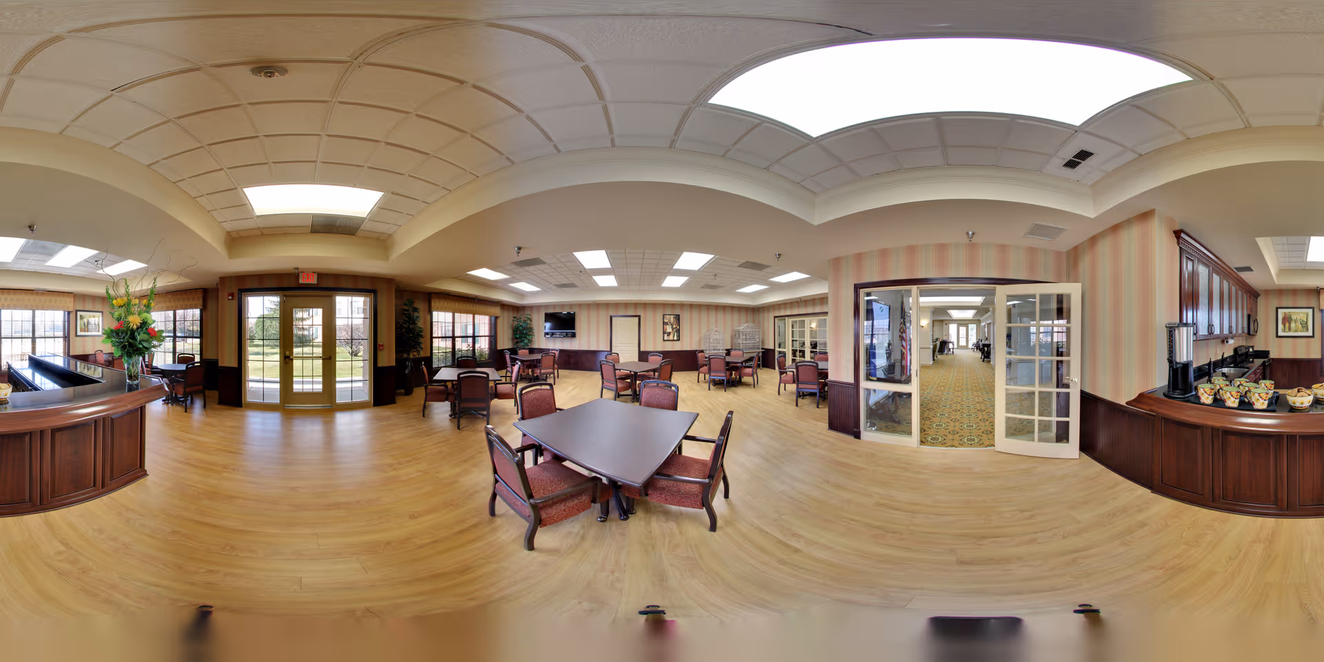 Spacious senior community dining and lounge area with multiple tables and chairs, a serving counter, and glass doors leading outside.