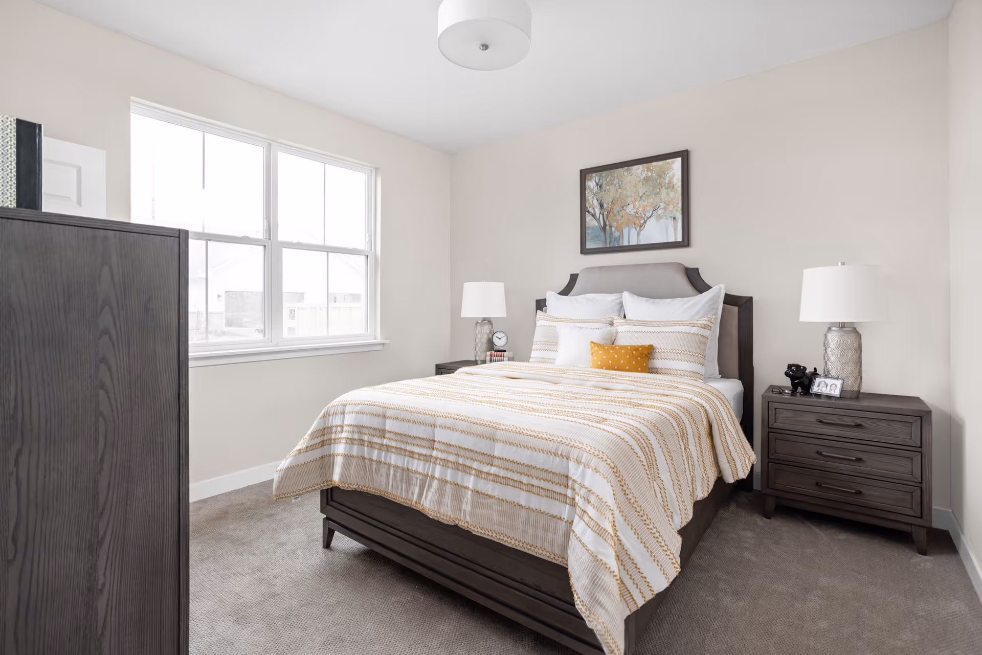 Bright furnished bedroom with a bed, two nightstands, dresser, window, and framed artwork on the wall.