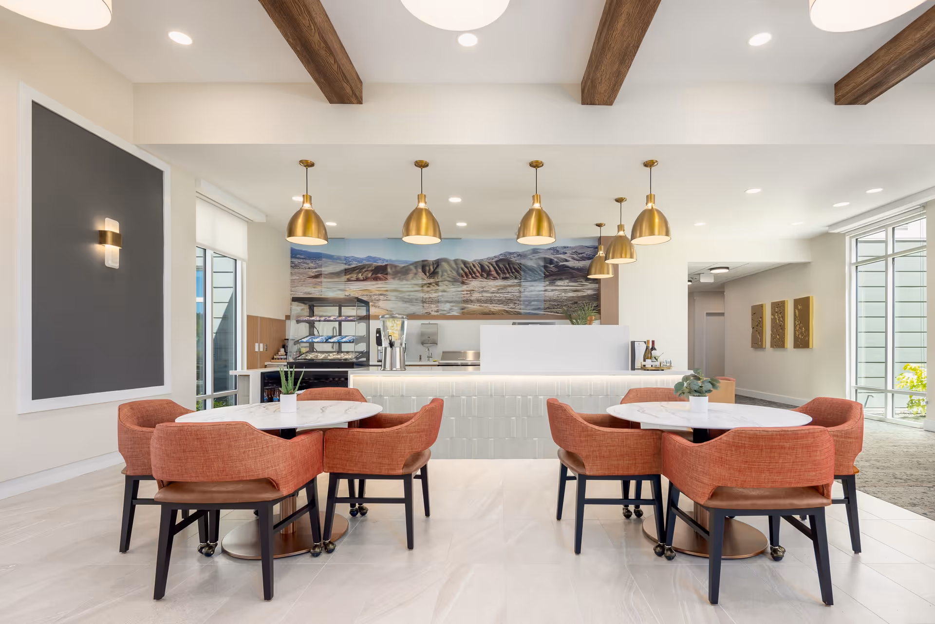 A bright dining area with two round white marble tables, each surrounded by four orange upholstered chairs with black legs. Above the tables hang six gold pendant lights. The back wall features a large scenic mural of a mountainous landscape. The ceiling has exposed wooden beams, and large windows allow natural light to fill the space.