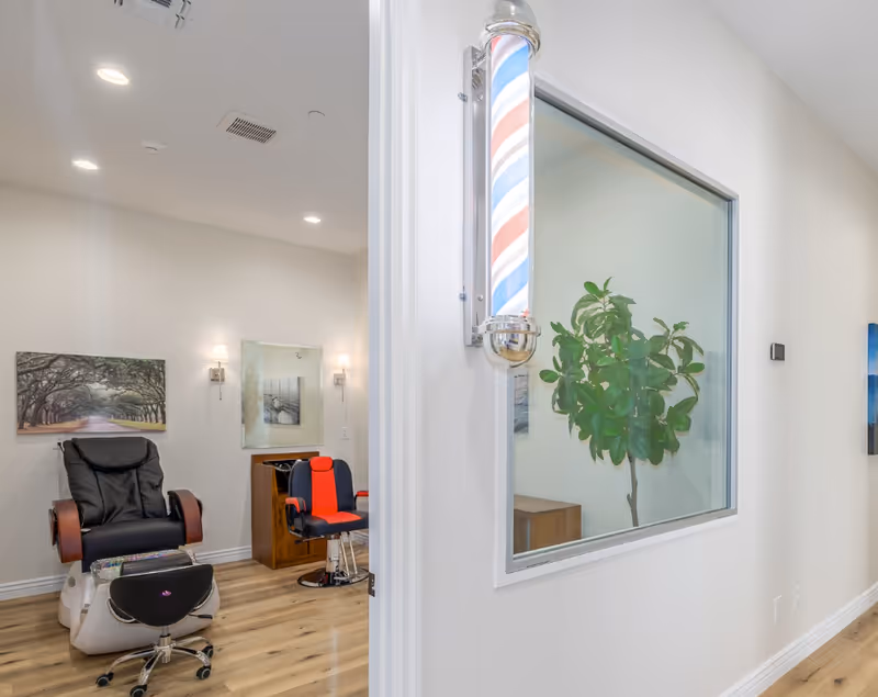 Interior salon space featuring a barber pole, pedicure chair, salon chair, and a potted plant seen through an interior window.