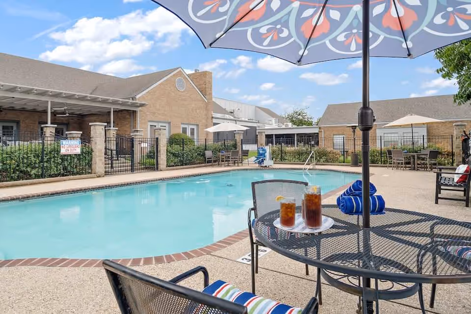 Outdoor swimming pool area at Morada Temple facility with a round metal table and chairs under a colorful umbrella. Two glasses of iced tea and rolled blue towels are on the table. The pool is surrounded by a concrete deck, patio furniture, and a black metal fence. The building with beige brick walls and multiple windows is visible in the background under a partly cloudy sky.