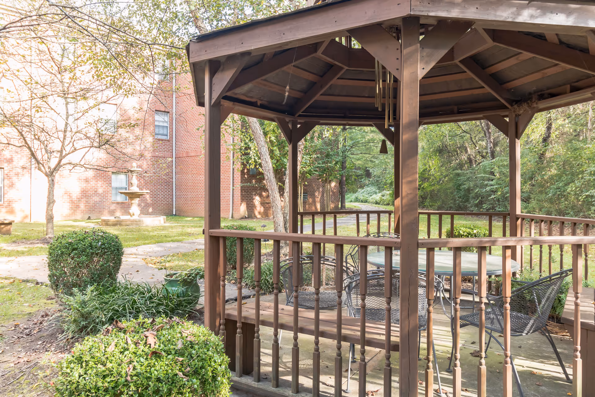 A wooden gazebo with a round glass table and metal chairs inside, situated in a garden area with green bushes, trees, and a brick building in the background. A stone fountain is visible near the building.