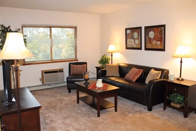 Well-lit living room with a black leather sofa and armchair, coffee table, TV stand, lamps, and a window.