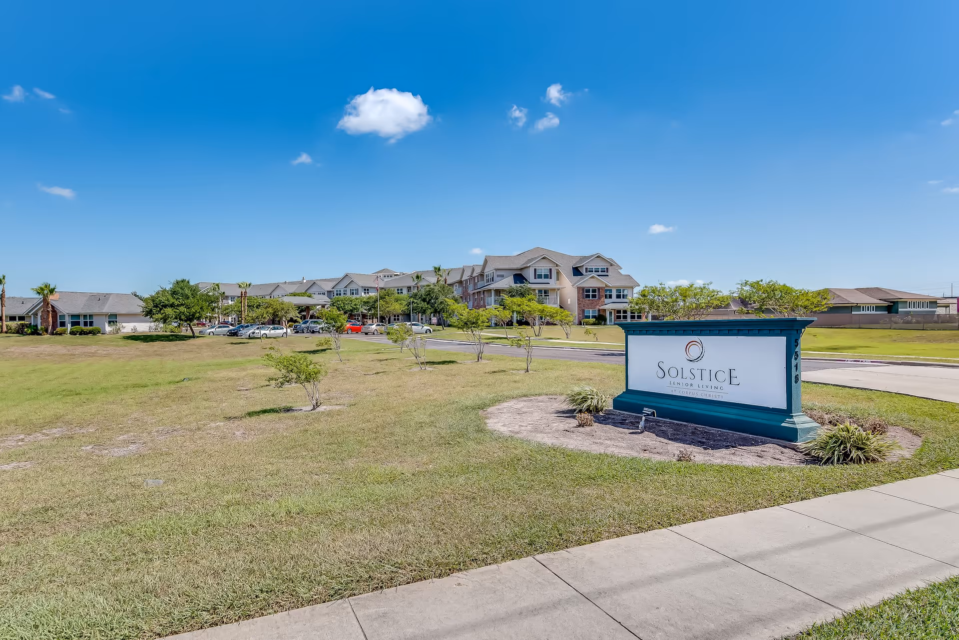 Exterior view of Solstice Senior Living at Corpus Christi facility on a sunny day with a clear blue sky. The image shows a large grassy area with small trees, a paved sidewalk, and a prominent sign displaying the facility's name. The senior living building is visible in the background with multiple windows and a parking area with several cars.