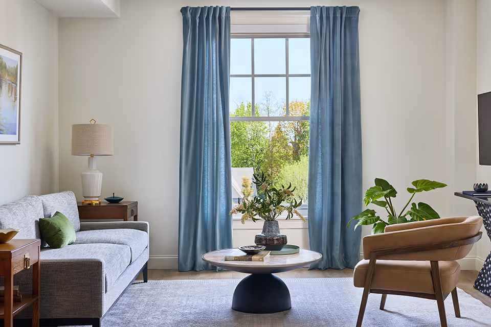 A bright living room with a large window framed by blue curtains. The room features a gray sofa with a green pillow, a wooden side table with a lamp, a round coffee table with a vase of flowers, a tan armchair, and a flat-screen TV mounted on the wall. The floor is covered with a light-colored rug.
