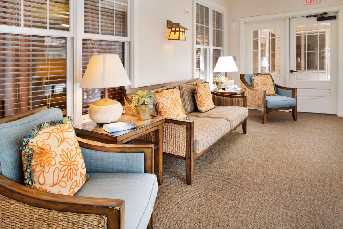 A cozy seating area with wooden framed chairs and a sofa featuring blue and striped cushions with orange floral pillows. Two side tables with lamps and a small plant are placed between the chairs and sofa. The room has beige carpet flooring, white walls, and large windows with white blinds. A white door with glass panels is visible in the background.