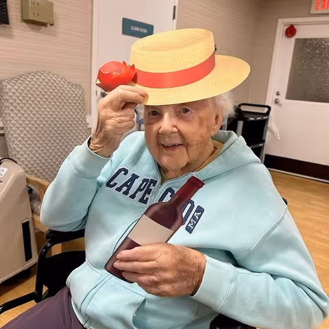 An elderly person sitting in a wheelchair indoors, wearing a light blue Cape Cod hoodie, holding a paper cutout of a wine bottle in one hand and a paper cutout of a straw hat with a red ribbon in the other hand, smiling at the camera. The background shows a beige wall, a door labeled 'Alpine Room,' and some chairs.