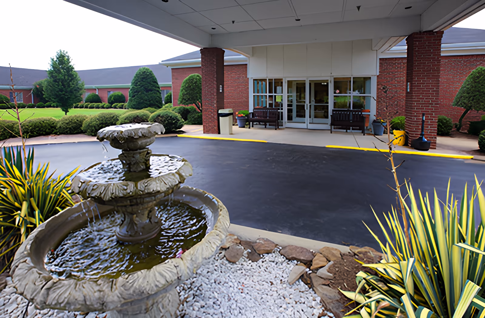 Entrance of Ahc West Tennessee Transitional Care facility showing a covered driveway with a three-tier stone water fountain in the foreground, surrounded by plants and landscaping. The building has red brick walls and glass double doors with benches on either side of the entrance.