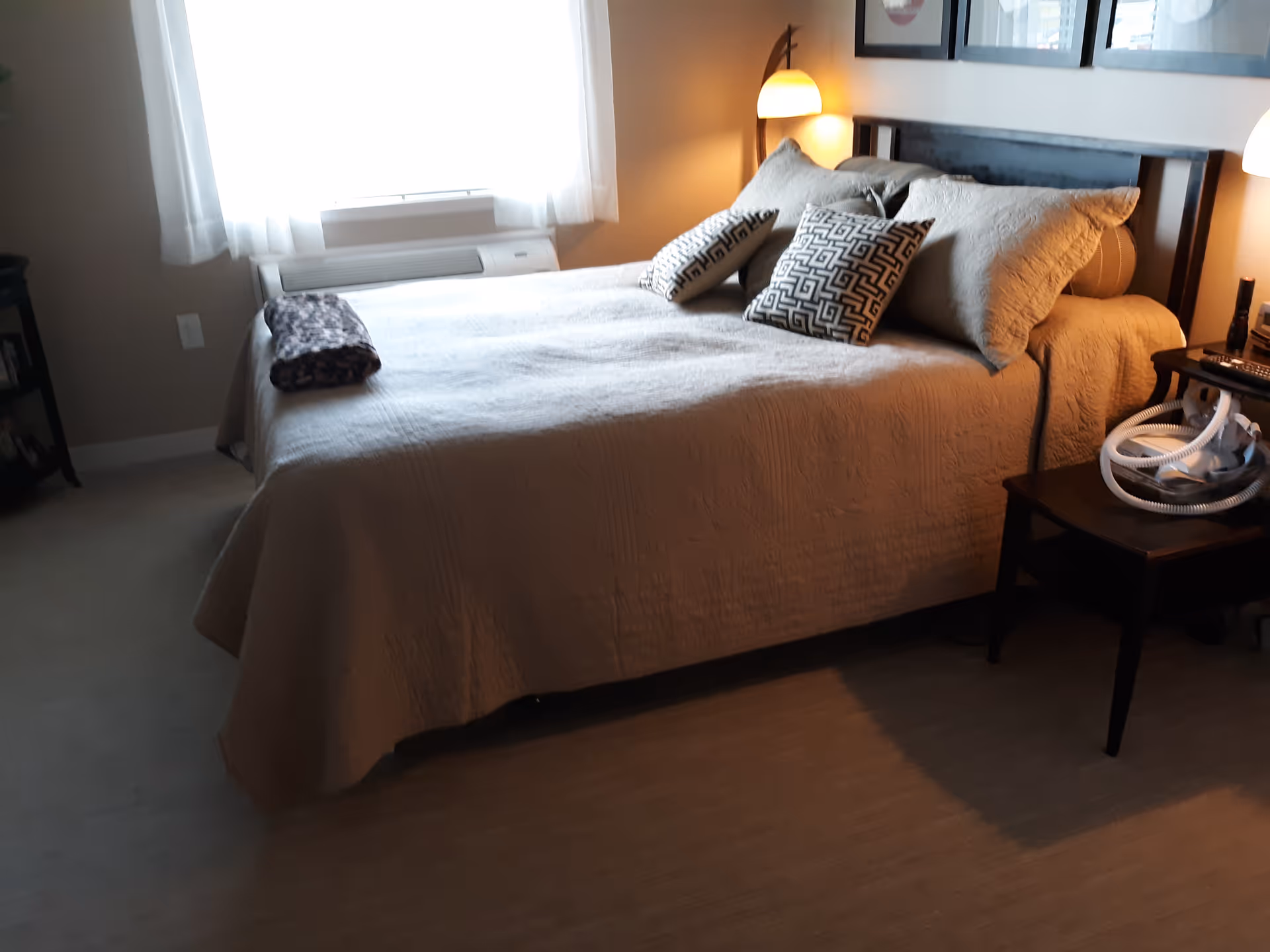 A cozy bedroom with a neatly made bed covered in a beige quilt and several decorative pillows. There is a rolled blanket at the foot of the bed. The room has a window with sheer white curtains allowing natural light to enter. On the right side of the bed, there is a nightstand with a lamp and a medical device with tubing. The walls are decorated with framed pictures.