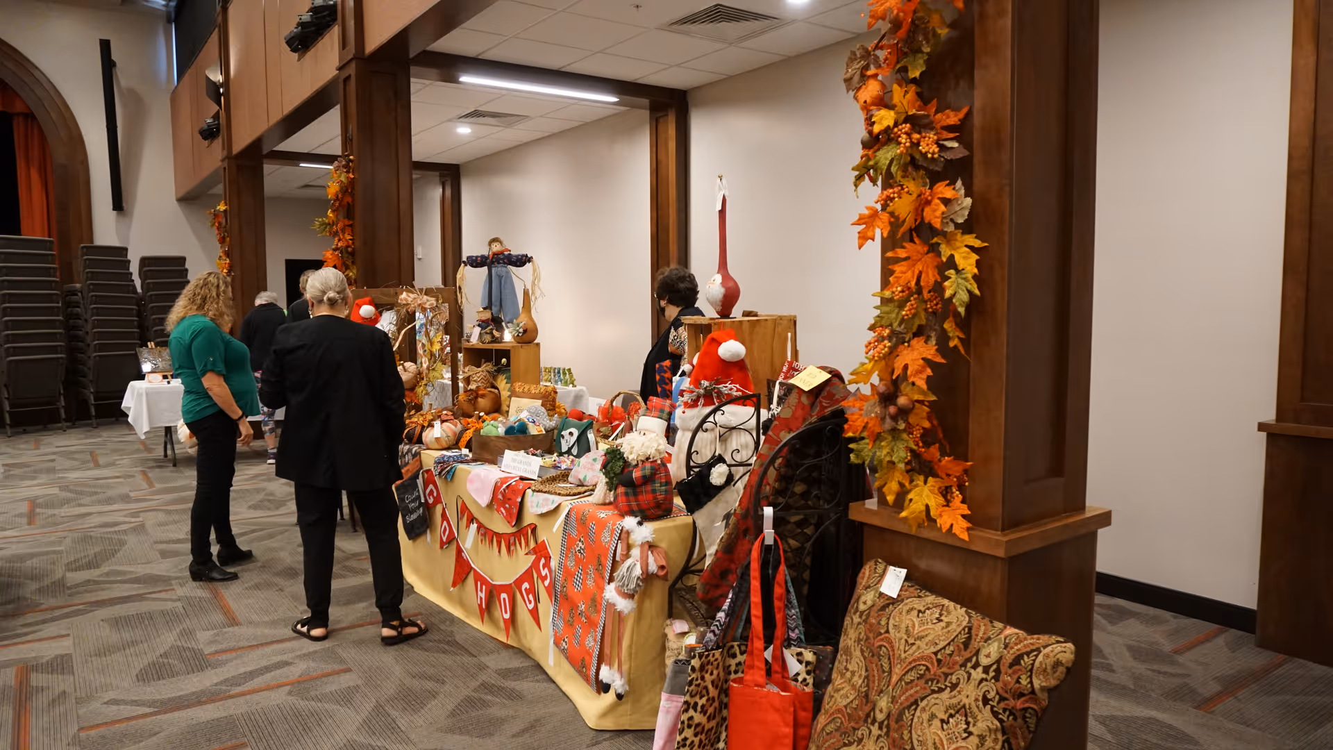 Indoor room with several people browsing a table covered with various handmade crafts and decorations. The table is draped with a yellow cloth and adorned with fall-themed garlands and items. The room has carpeted floors, wooden pillars decorated with autumn leaves, and stacked chairs in the background.