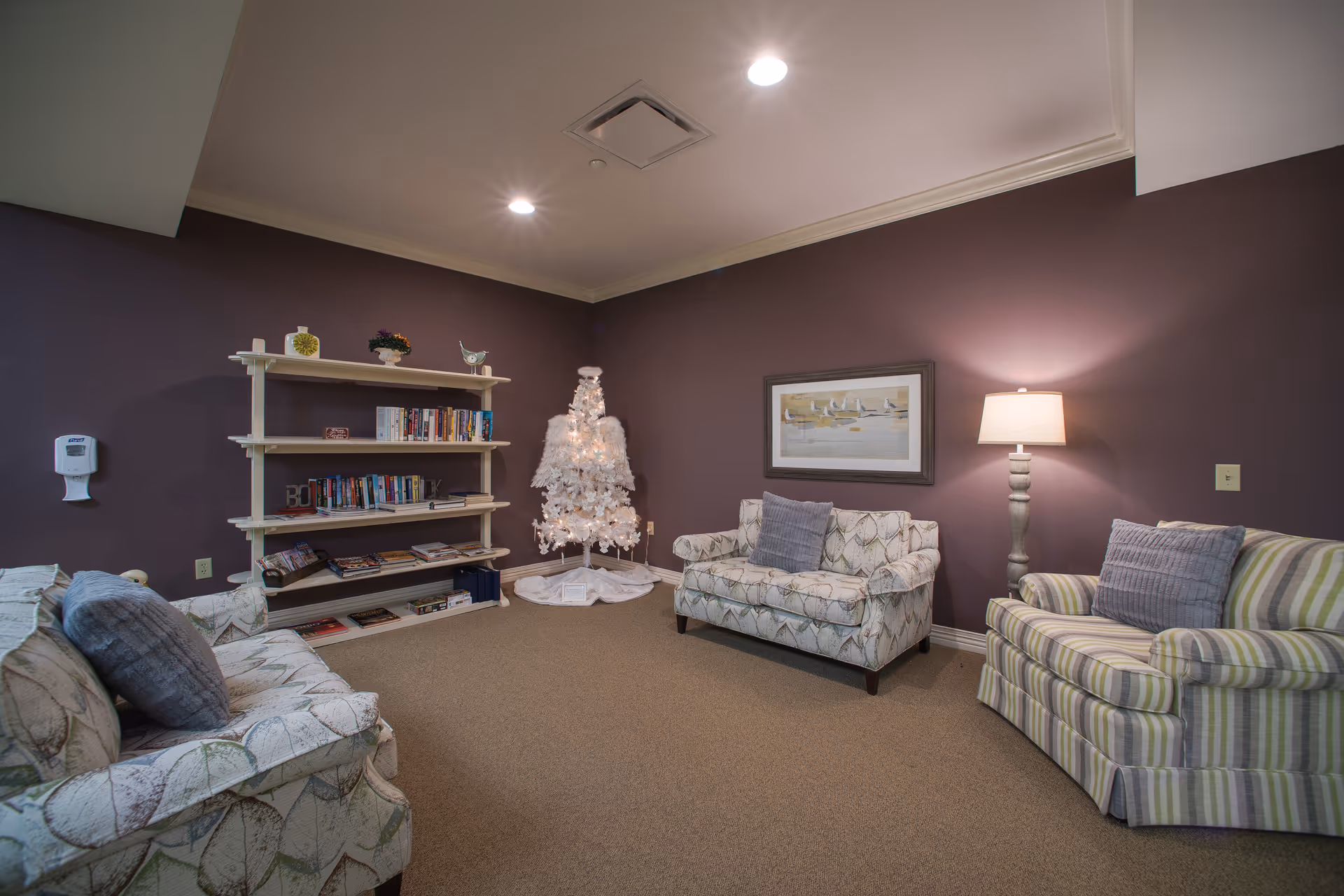 A cozy living room with three patterned armchairs arranged around a beige carpeted floor. A white Christmas tree with lights stands in the corner next to a white bookshelf filled with books and decorative items. A floor lamp with a beige shade illuminates the room, which has purple walls and a framed painting of sailboats.