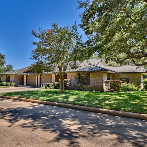 Single-story brick building with a metal roof surrounded by green grass, bushes, and trees under a clear blue sky.