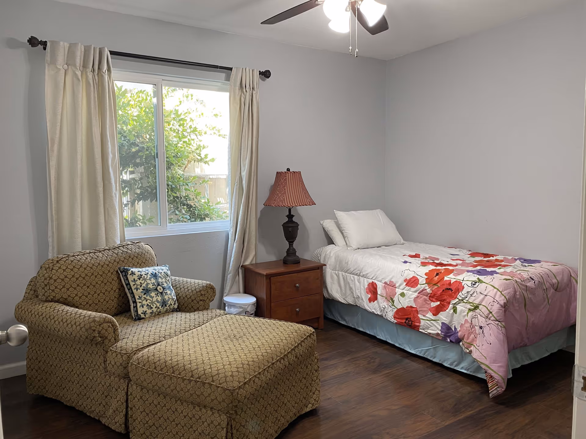 A small bedroom with a single bed covered with a floral comforter, a wooden nightstand with a lamp, a cushioned armchair with an ottoman, and a window with cream-colored curtains showing greenery outside.