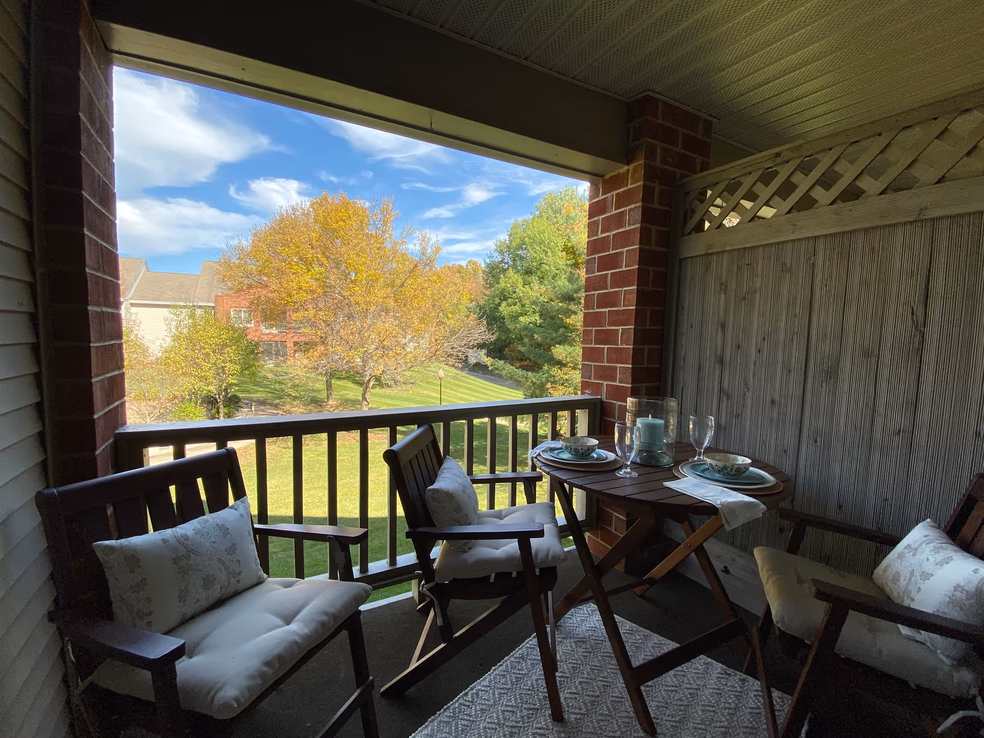 Covered balcony with cushioned wooden chairs and a small table set for two overlooking a grassy courtyard and trees.