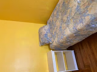 A small bedroom with yellow walls and wooden flooring. The room contains a single bed with a floral-patterned bedspread and a white bedside table with two shelves next to the bed.