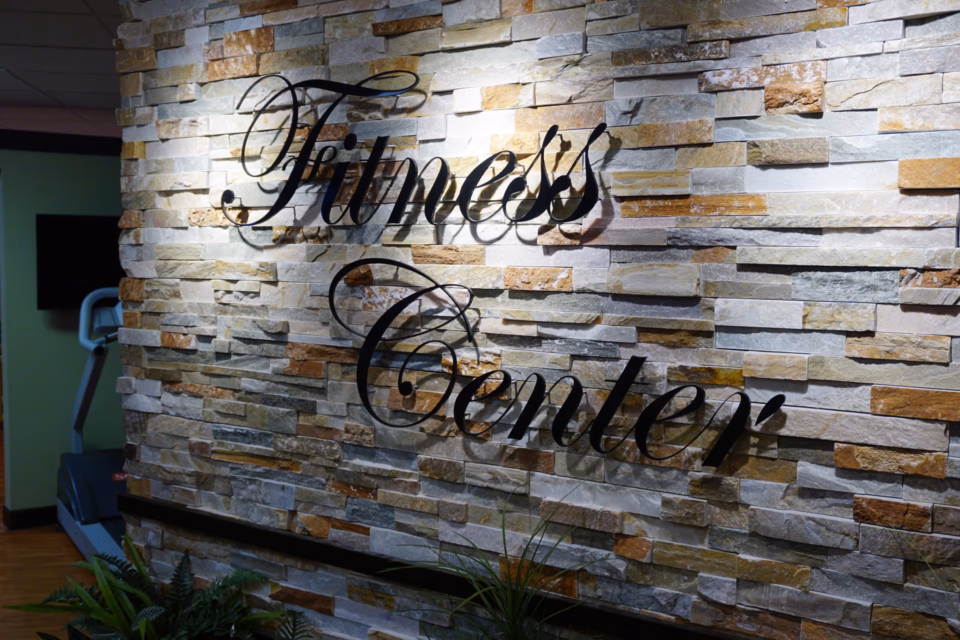 A stone wall with the words 'Fitness Center' mounted on it in elegant black script. There is a treadmill partially visible to the left and some green plants at the bottom of the wall.