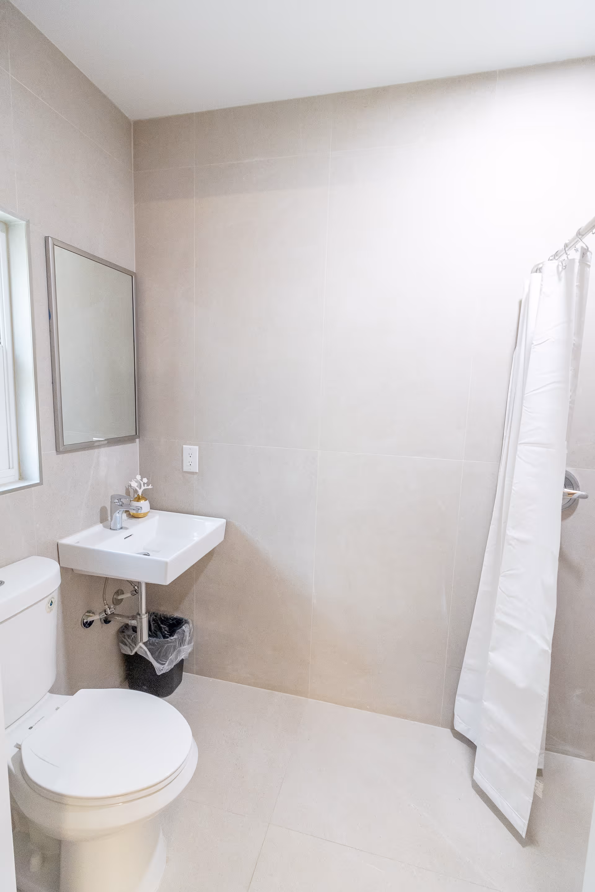 Compact bathroom with a toilet, wall-mounted sink and mirror, small trash can, and a shower with a white curtain.