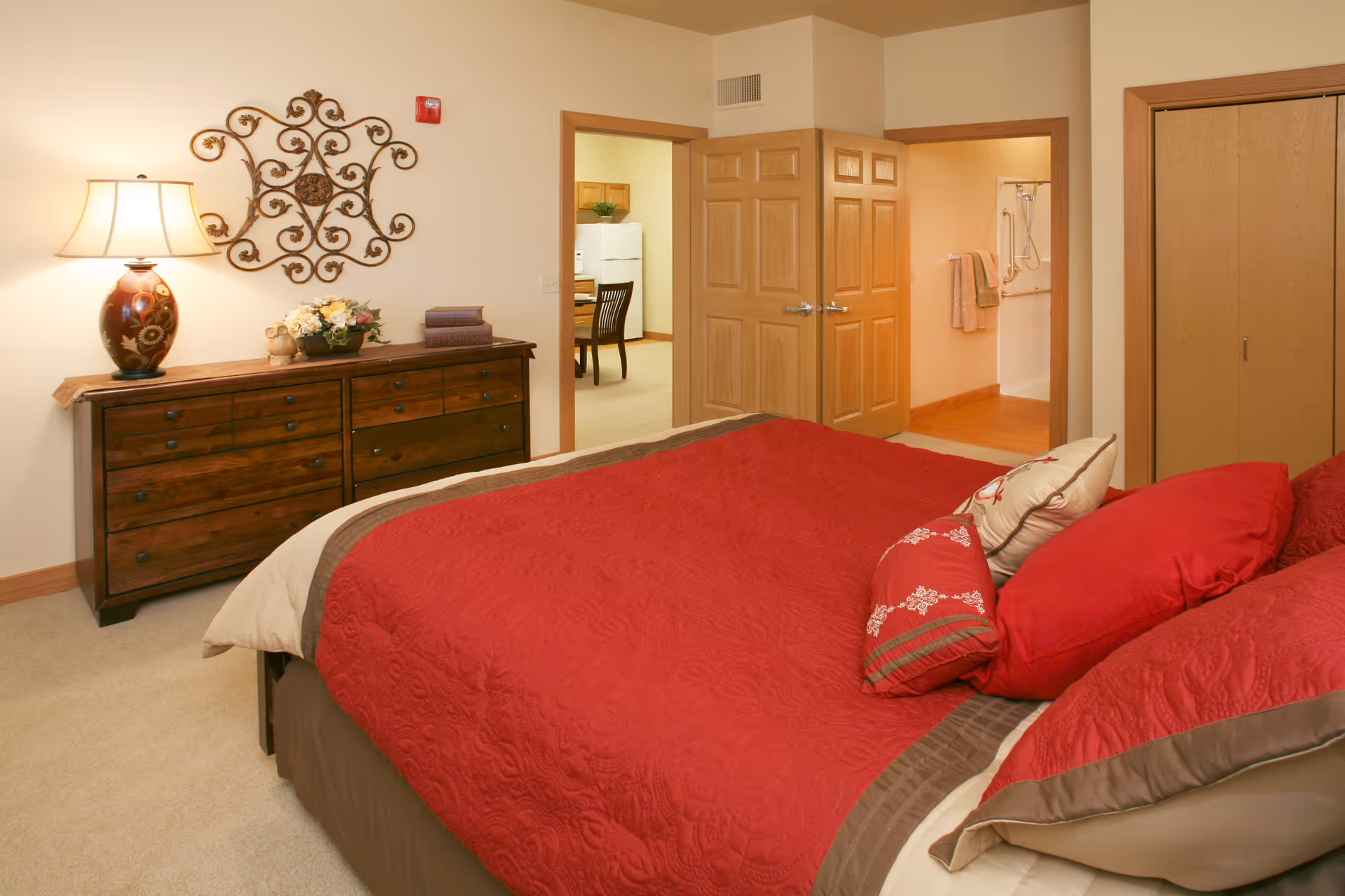 A bedroom with a bed covered in red bedding and multiple pillows. To the left is a wooden dresser with a decorative lamp, flowers, and books on top. Above the dresser is a decorative metal wall art. The room has beige walls and carpet. Two open doors reveal a kitchen area with a refrigerator and a bathroom with a shower and towels.