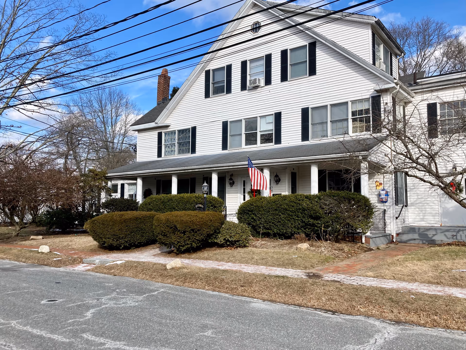Exterior view of a large white multi-story building with black shutters and a front porch supported by white columns. There is an American flag displayed on the porch, surrounded by well-maintained bushes and a brick walkway leading to the entrance. The sky is clear with some clouds, and leafless trees are visible in the background.