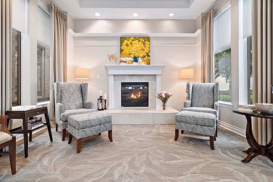 A cozy living room with two patterned armchairs and a matching ottoman facing a lit fireplace. The fireplace mantel is decorated with a bouquet of flowers and a painting of yellow flowers above it. There are large windows on both sides with beige curtains and roller shades, and side tables with books and decorative items.
