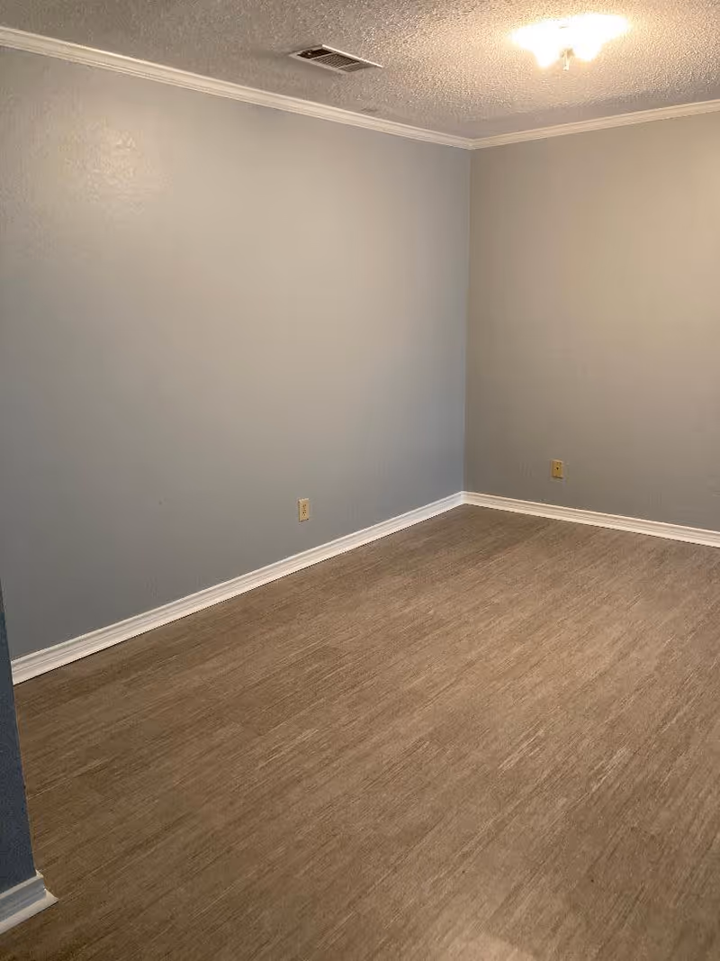 Empty room with light gray walls, wood-like flooring, white baseboards, a ceiling light fixture, and two electrical outlets on the walls.