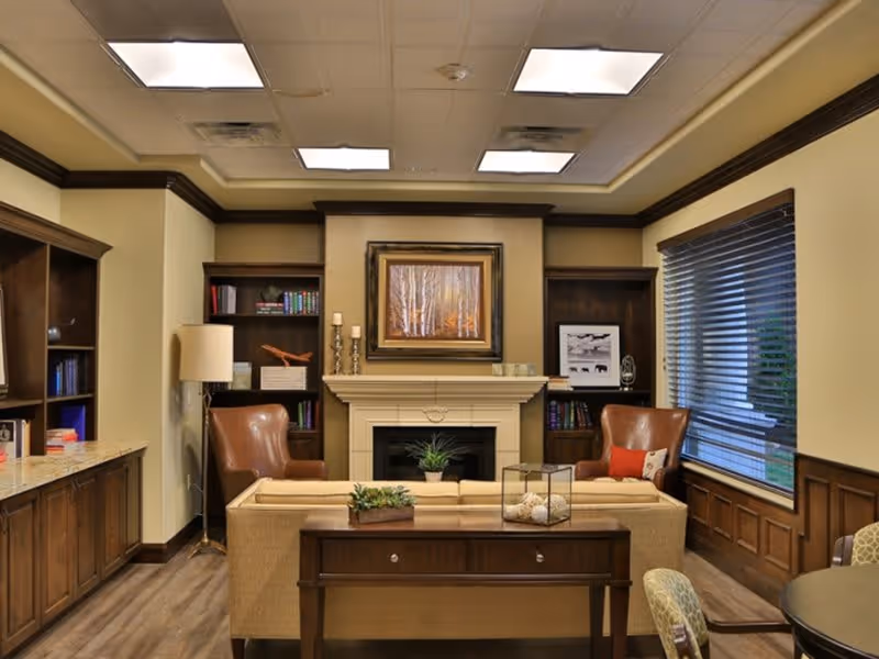 A cozy living room area with a beige sofa facing a fireplace. Two brown leather armchairs flank the fireplace, which has a framed painting above it. Built-in wooden bookshelves with books and decorative items are on either side of the fireplace. A large window with blinds is on the right wall, and a wooden console table with decorative plants is behind the sofa. The room has warm lighting and a wood floor.