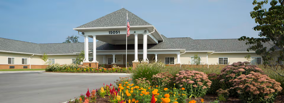 Front entrance of a single-story health and rehab building with a covered portico, an American flag, and colorful flowerbeds in the foreground.