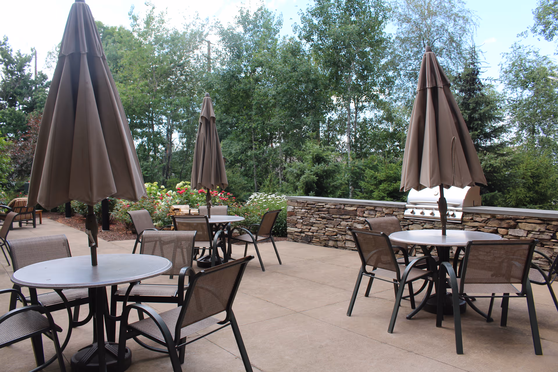 Outdoor patio area with round tables and chairs, each table having a closed brown umbrella. There is a stone wall with a built-in grill in the background and trees and greenery surrounding the patio.