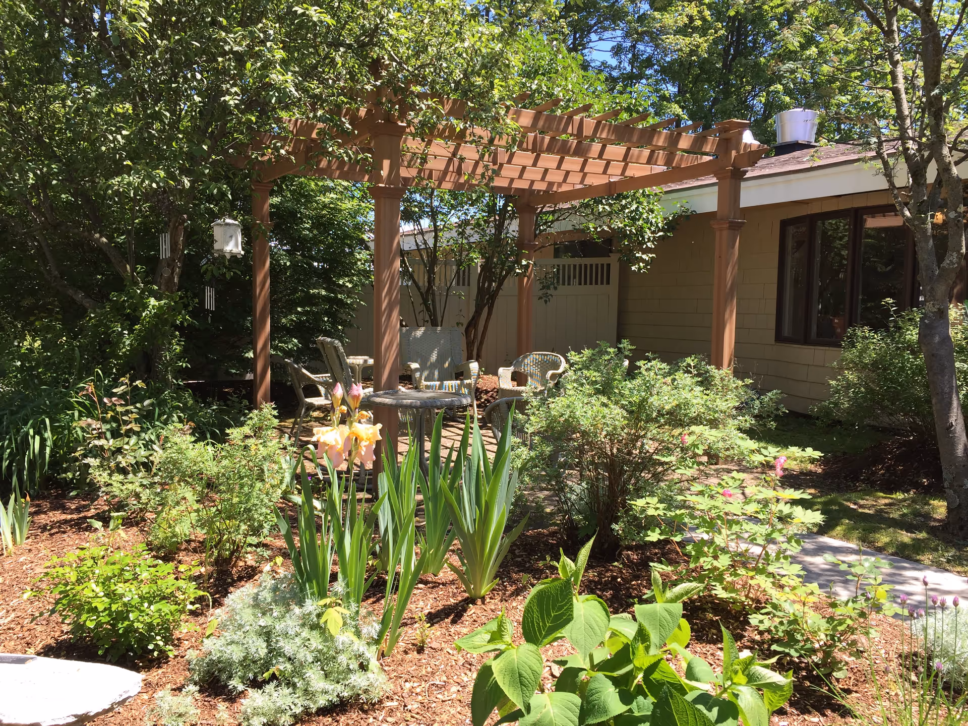 A sunny outdoor garden area with a wooden pergola providing partial shade over a set of wicker chairs and a small table. The garden is lush with various green plants, bushes, and blooming flowers, surrounded by trees and a building with beige siding and windows in the background.